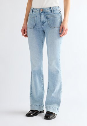 Jean bleu clair taille haute avec poches plaquées à l'avant, porté par une personne en haut blanc et mocassins noirs sur fond uni.