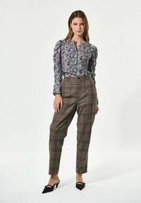 Bluse mit floralem Muster in Hellblau und Puffärmeln; braune Karo-Hose mit hohem Bund; schwarze spitze High Heels; neutraler Hintergrund.