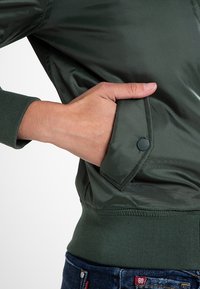 Oliwkowa bomberka z boczną kieszenią, prążkowanymi mankietami i gładką, błyszczącą teksturą. Posiada detal z zatrzaskiem na kieszeni.