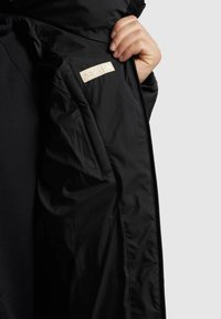 Sort jakke indvendig med en glat stofstruktur, lynlåslukning og et lille cremefarvet mærke med præget detaljer.