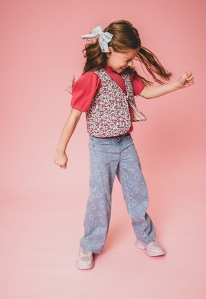 Bloemenpatroon vest over een rode geribde t-shirt met korte mouw, gecombineerd met lichtblauwe spijkerbroek met hartjespatroon en roze sneakers.