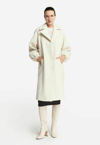 Vit ullblandad kappa med snibbat krage, överdimensionerade ärmar och framfickor, bärs över en svart kjol och beige klackstövlar.