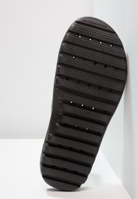 Musta kumipohja kengästä, jossa on vaakasuoria uria ja pieniä pyöreitä reikiä, asetettuna valkoisen seinän ja lattian kulmaa vasten.