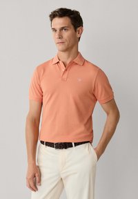Homme portant un polo à manches courtes de couleur saumon, un pantalon blanc et une ceinture tressée foncée, debout avec une main dans la poche.