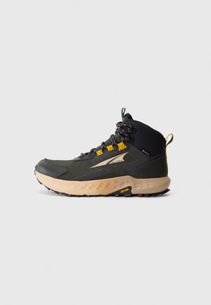 Hiking boot with a black and olive green upper, featuring a textured fabric and yellow accents. Thick, tan sole with Vibram logo.