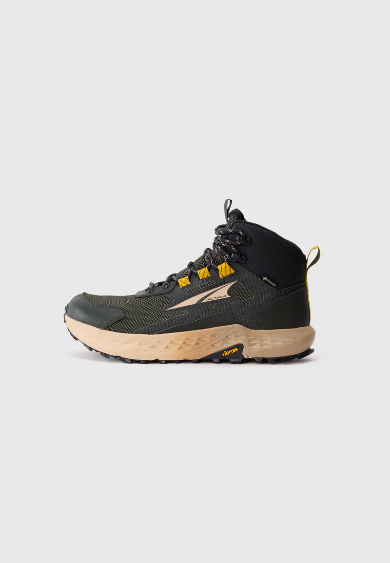 Fjellstøvel med en svart og olivengrønn overdel, som har et teksturert stoff og gule detaljer. Tykk, beige såle med Vibram-logo.
