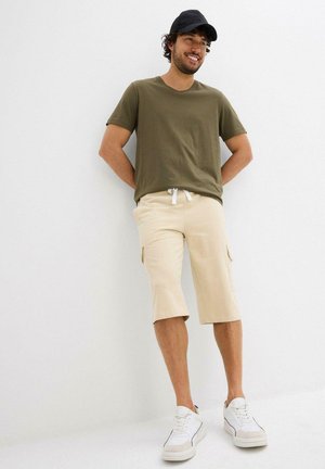 T-shirt khaki abbinata a pantaloni cargo beige con tasche laterali, completata da sneakers bianche con accenti grigi. Design casual.