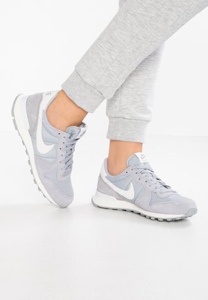 Osoba nosząca jasnoszare buty Nike z białym logo i pasujące szare spodnie dresowe, stojąca na białej podłodze.