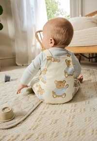 Salopette en coton écru avec imprimés d'ours en peluche ; sous-vêtement rayé bleu clair ; tapis crème doux et texturé en dessous. Jouets à proximité.