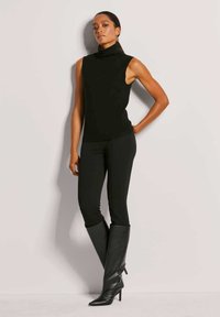 Schwarzes ärmelloses Rollkragenoberteil kombiniert mit schwarzen enganliegenden Hosen und kniehohen Stiletto-Stiefeln. Glatter Stoff mit einem eleganten, figurbetonten Design.