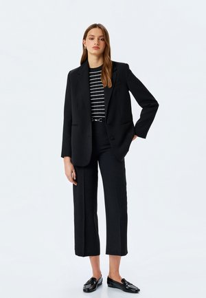 Blazer negro con solapas tipo muesca, llevado sobre una camisa a rayas en blanco y negro, combinado con pantalones cropped negros y mocasines negros brillantes.