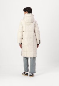 Lang, lys beige pufferjakke med hette, med horisontale sømdetaljer og sideslister. Kombinert med lyseblå jeans og svarte joggesko.