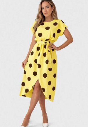 Femme portant une robe midi jaune à gros pois noirs, ceinture nouée à la taille, manches courtes, fente à l’avant et talons blancs.