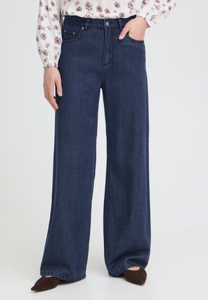 Femme portant un jean large taille haute bleu foncé avec un chemisier à manches longues à motifs floraux, sa main gauche dans la poche et des chaussures marron pointues.