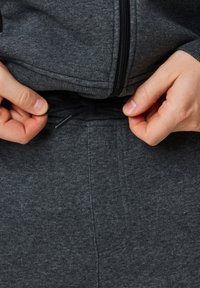 Grå fleece-tröja med dragkedja, en dragsko vid midjan och textilstruktur. Händer som justerar midjebandet syns.