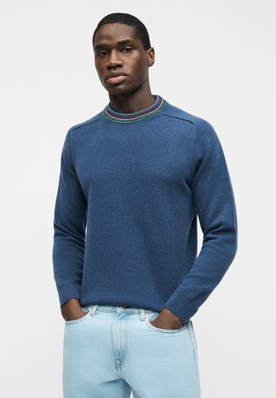 Homme à la peau foncée portant un pull bleu avec un col rayé multicolore et un jean bleu clair, debout les mains dans les poches.