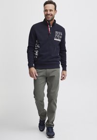 Felpa navy con mezza zip, caratterizzata da testo grafico bianco e rosso, abbinata a pantaloni verde chiaro e sneakers navy.