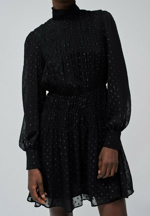 Czarna, przezroczysta sukienka z długim rękawem i wysokim kołnierzem, z delikatnym kwiatowym wzorem, marszczonymi talią i mankietami, noszona przez osobę na jednolitym tle.