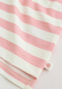 Mekani materijal s vodoravnim ružičastim i bijelim prugama, sa sitnom bijelom oznakom na kojoj je ružičasti cvjetni simbol prišiven blizu ruba.