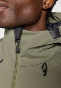 Olívovo zelená bunda má vysoký golier, zips a nastaviteľné šnúrky. Materiál má hladkú textúru s matným povrchom.