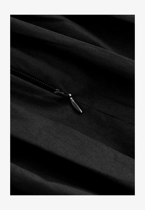 Zwarte stof met subtiele textuur, voorzien van een ritssluiting. De rits heeft een metalen trekker, geplaatst tegen een gladde, hangende stof.