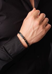Bracciale in metallo nero con anelli intrecciati, caratterizzato da un pezzo accentato lucido. Indossato attorno al polso di una mano su una camicia scura.