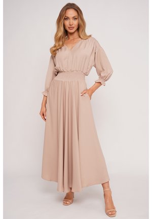 Kobieta ubrana w długą beżową sukienkę z taliowanym pasem i rękawami o długości do łokcia z bufkami, pozująca z jedną ręką w kieszeni, nosząca neutralne szpilki.