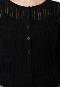 Schwarzer, gestrickter Cardigan mit vertikalen Rippen und kleinen runden Knöpfen in der Mitte, getragen am oberen Rumpf und Halsbereich.