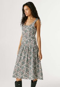 Frau trägt ein ärmelloses, knieumspielendes Kleid mit Blumenmuster und geraffter Taille sowie schwarze Stiefel bis zur Mitte der Wade, steht vor schlichtem Hintergrund.