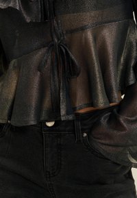 Schwarze, schimmernde Bluse mit einem Rüschensaum und Bindebändern am Dekolleté; kombiniert mit schwarzen Denim-Jeans, die einen Knopf- und Taschenakzent zeigen.