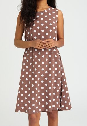 Ærmeløs brun kjole med hvide polkaprikker, knælængde, båret af en person med hænderne blidt foldet foran.