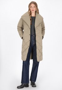 Beige steppelt hosszú kabát, amely nagy gallérral rendelkezik, zapfogó gombokkal és oldalsó zsebekkel készült, sötétkék trapéznadrággal és fekete bokacsizmával párosítva.