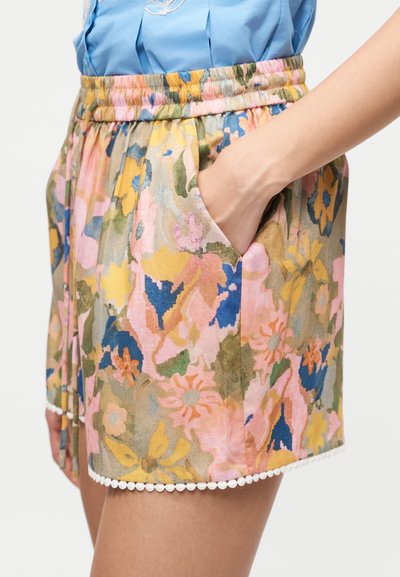 Γυναίκα που φοράει ψηλόμεσες φλοράλ σορτς με ελαστική μέση και λευκό πομπόν περίγραμμα, το χέρι της ακουμπάει στην δεξιά τσέπη, σε συνδυασμό με μπλε πουκάμισο.