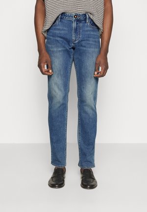 Homme portant un jean bleu et des chaussures en cuir noir, debout devant un fond uni, les mains détendues le long du corps.