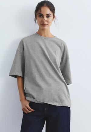 Vrouw draagt losse grijze T-shirt met korte mouwen en donkerblauwe spijkerbroek, staand tegen een effen lichte achtergrond met hand in de zak.