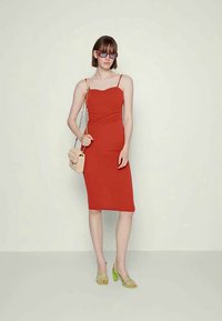Vestido midi justo vermelho com alças finas, detalhes texturizados no corpete, combinado com sandálias verdes e uma pequena bolsa bege.