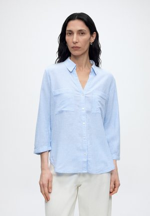 Femme aux longs cheveux foncés portant une chemise rayée bleu clair à boutons avec des poches et un pantalon blanc sur un fond uni.