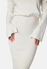 Abito bianco a costine con maniche lunghe e ampi polsini, caratterizzato da una silhouette aderente e una sottile texture per un ulteriore interesse visivo.