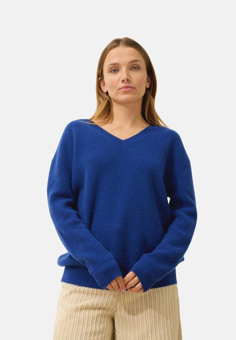 Blå V-ringad tröja i strukturerad stickad kvalitet, med ribbade mudd och nederkant. Modellen har på sig krämfärgade ribbade byxor.