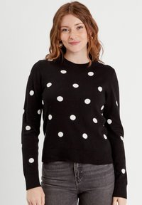 Zwarte trui met witte stippen, ronde halslijn, lange mouwen en een iets cropped pasvorm. Zachte textuur met geribbelde manchetten en zoom.