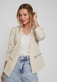 Sacou bej cu o croială ajustată, umeri structurați, două buzunare laterale cu fermoar, asortat cu un top alb și blugi cu talie înaltă.