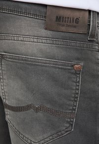 Jeans en denim gris foncé avec une étiquette en cuir emboutie "MUSTANG True Denim", design à cinq poches et détails de couture contrastés.
