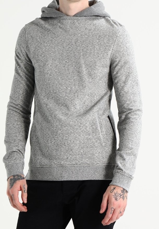 Homme portant un sweat à capuche gris clair à manches longues et un pantalon noir, montrant des mains et des avant-bras tatoués.