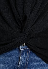 Svart stickad topp snörts vid midjan som blottar en midja av blå denim med en metallknapp. Texturen verkar mjuk och lätt.