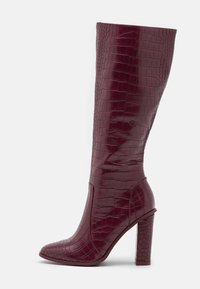 Bottes bordeaux montant jusqu'au genou avec une finition brillante embossée effet croco, bout pointu et talon épais texturé. Dotées d'un design épuré et d'accents de couture.