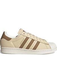 Beige Adidas Superstar sneaker with brown stripes, textured heel tab, white rubber shell toe, and flat sole, side view on white background.