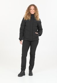 Schwarze Steppjacke mit Reißverschluss vorne und seitlichen Taschen, kombiniert mit schwarzen Hosen und robusten schwarzen Stiefeln, mit taillierter Passform und hohem Kragen.