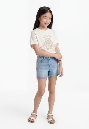 Jeune fille aux longs cheveux foncés porte un t-shirt blanc Versace, un short en jean bleu clair et des sandales blanches, debout les bras croisés, souriante.