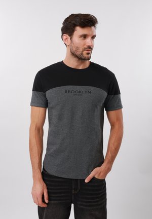 Muži s bradou v čiernom a sivom tričku "Brooklyn New York", pozerajúci sa do strany, s jednou rukou v džínovej vrecku.