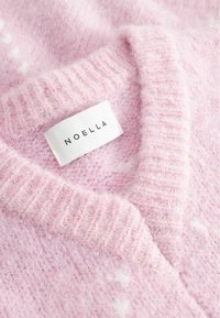 Helles rosa gestricktes Sweatshirt mit einem gerippten Kragen und ausgeprägter Textur, mit einem weißen Etikett, das den Markennamen "NOELLA" zeigt.